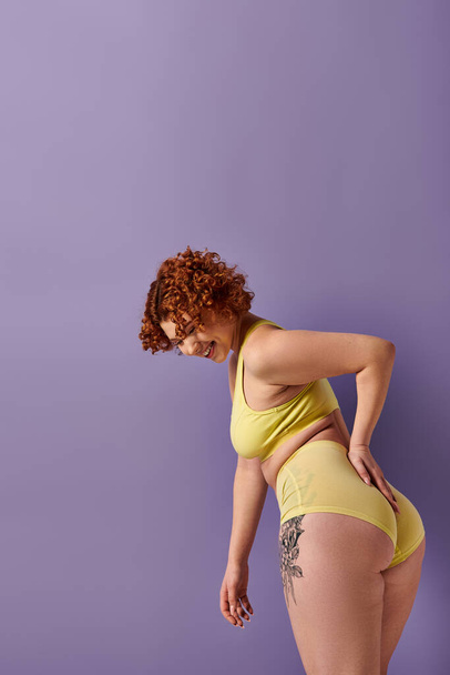 Curvy redhead woman striking a pose in yellow bikini against vibrant purple backdrop. - Foto, imagen