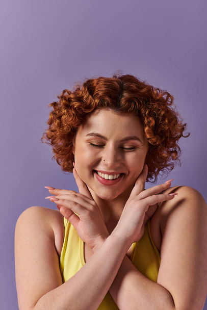 A young, curvy redhead woman in yellow underwear strikes a playful pose, hands on face, against a purple background. - Foto, Bild