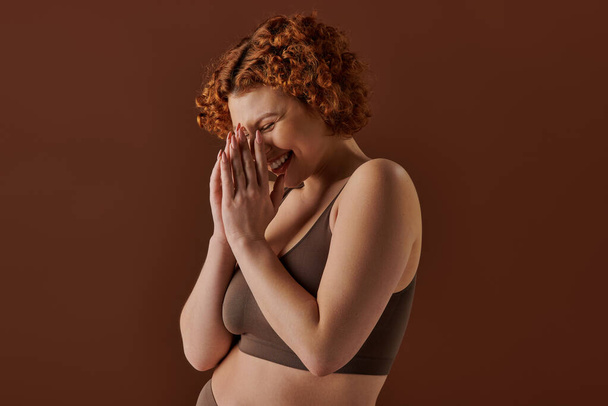 Curvy redhead woman in brown bikini, hands covering face delicately. - Photo, image