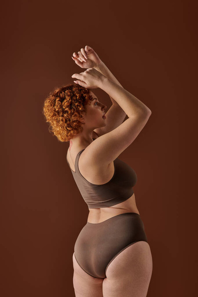 Young curvy redhead woman in brown bikini striking a pose with hands raised. - Foto, Bild