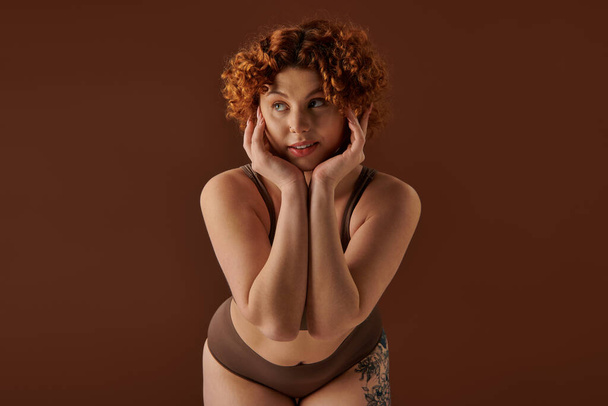 A curvy redhead woman showcases elegance and sensuality in lingerie against a brown background. - Foto, Imagem