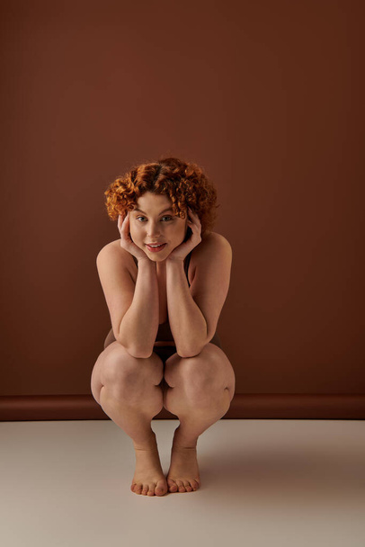 A young, curvy redhead woman crouches gracefully on the floor in her underwear against a brown backdrop. - Foto, afbeelding