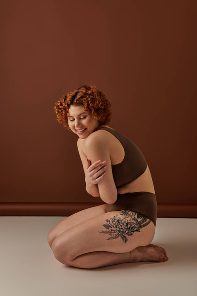 A young, curvy redhead woman sits gracefully on the floor in a cozy brown room. - Foto, Imagem