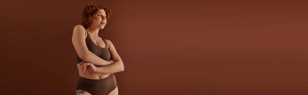 A young, curvy redhead woman in a bikini exudes confidence in front of a brown background. - Foto, Imagen