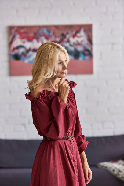 Kypsä nainen silmiinpistävä punainen mekko seisoo sulavasti viihtyisässä olohuoneessa. - Valokuva, kuva