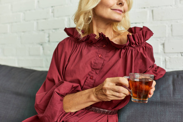 volwassen vrouw in stijlvolle jurk genieten van een moment van ontspanning met een glas thee op een bank. - Foto, afbeelding
