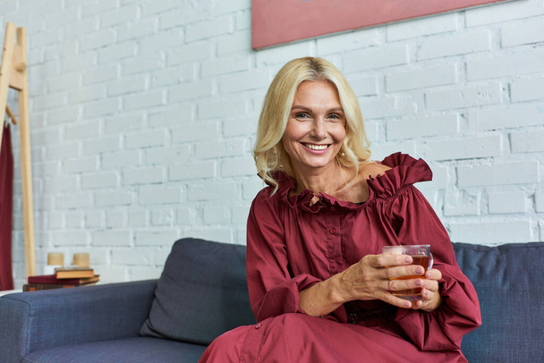 Reife Frau in stilvollem Kleid sitzt auf einer Couch und hält eine Tasse Tee in der Hand. - Foto, Bild