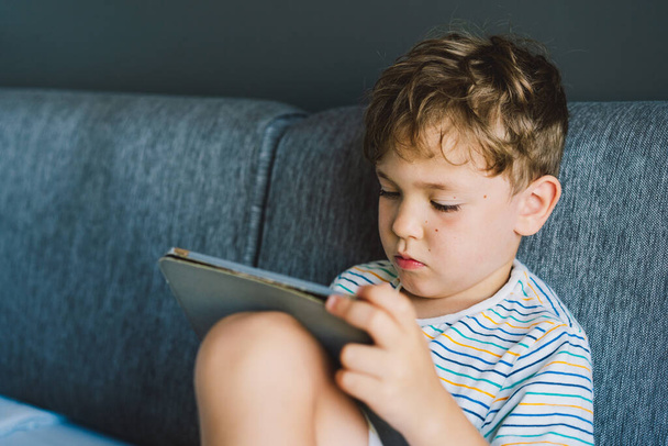 Ein kleiner Junge sitzt auf einer grauen Couch und spielt konzentriert Tablet. Er trägt ein T-Shirt und wirkt bequem und in seine Tätigkeit vertieft, was auf einen ruhigen und friedlichen Nachmittag drinnen hindeutet. - Foto, Bild