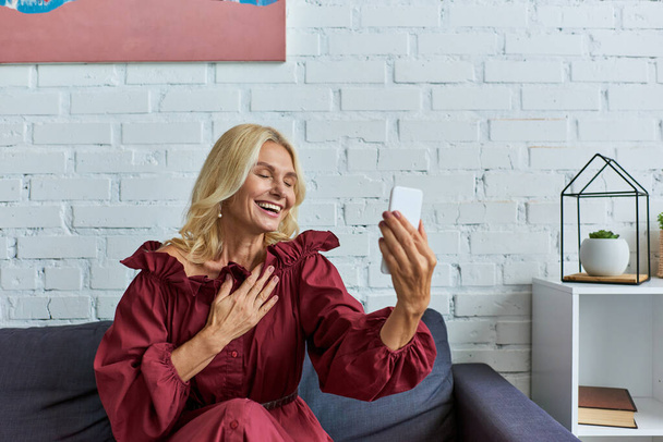 Mature woman in stylish dress, holding cell phone on couch. - Photo, Image