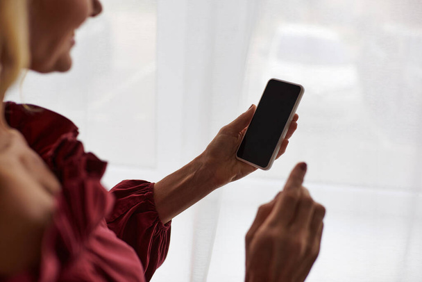 Mujer madura en una camisa roja con estilo sostiene un teléfono celular en casa. - Foto, imagen