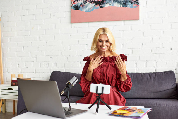 Ώριμη γυναίκα σε κομψό κόκκινο φόρεμα δημιουργώντας ένα podcast για γυναικεία ομορφιά μπροστά από ένα φορητό υπολογιστή. - Φωτογραφία, εικόνα