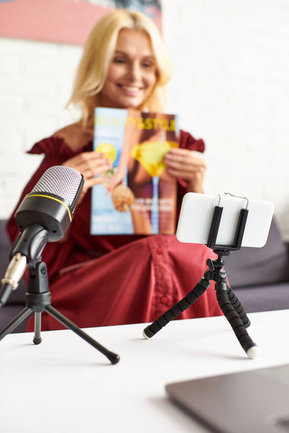 Egy érett, elegáns nő, piros elegáns ruhában, aki egy mikrofon előtt ül, kezében egy magazinnal.. - Fotó, kép