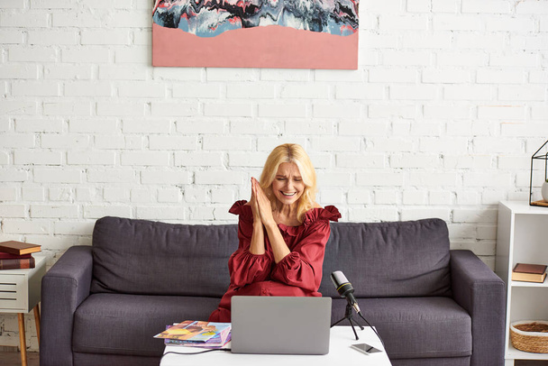 Kırmızı şık elbiseli olgun bir kadın dizüstü bilgisayarın önünde koltukta oturuyor ve kadın güzelliği üzerine podcast yapıyor.. - Fotoğraf, Görsel