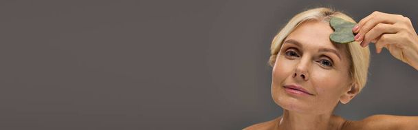 Gorgeous mature woman using gua sha on a gray backdrop. - Photo, Image