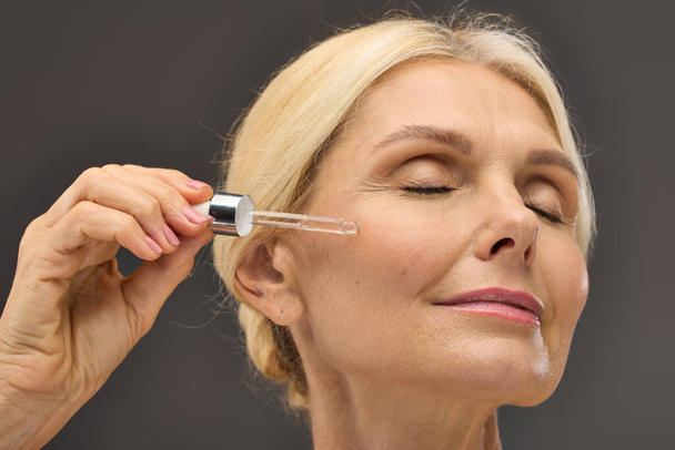 Gorgeous mature woman applying serum on a gray backdrop. - Photo, Image