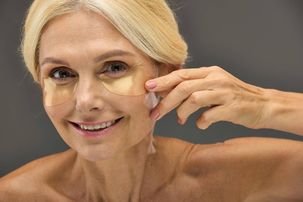 Dojrzała kobieta prezentująca opaski na oczy podczas stosowania pielęgnacji skóry. - Zdjęcie, obraz