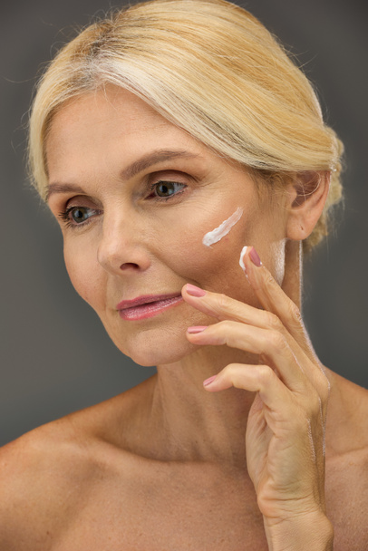 Femme mûre avec crème sur le visage pose sur un fond gris. - Photo, image