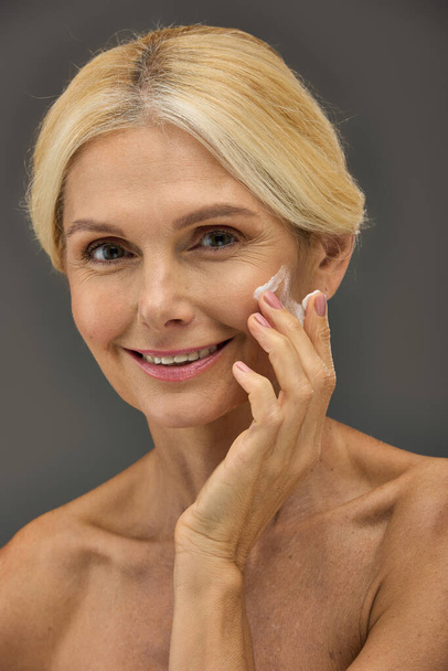 Mujer madura sonriendo y aplicando crema en su cara sobre un fondo gris. - Foto, Imagen