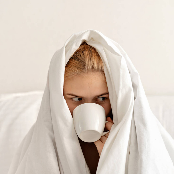 Ein junges Teenager-Mädchen in weiße Decke gehüllt sitzt auf dem Bett, hält eine Tasse in der Hand und trinkt erfrischenden Kaffee. Schwer aufwachendes Morgenkonzept, Schlafmangel und ein gesunder Lebensstil für Teenager. - Foto, Bild