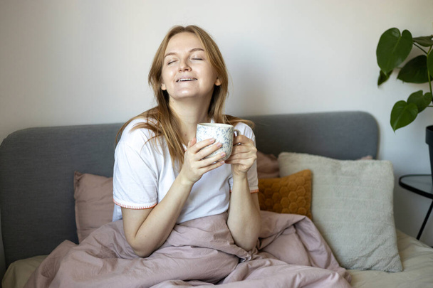 Retrato de una guapa joven rubia sosteniendo una taza con bebida caliente. Mujer relajante en la cama acogedora en casa. Bebidas y concepto de ocio - Foto, Imagen