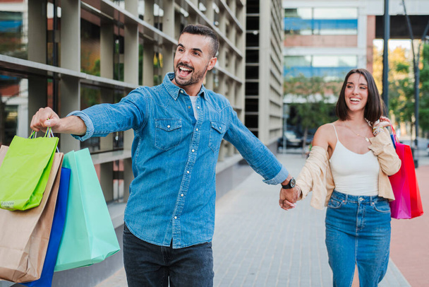 A boltos fickó fut és húzza a barátnője karját, elmegy egy butikba. Fiatal felnőtt vásárlók, akik gyorsan sétálnak a plázába bevásárlószatyrokat cipelve, hogy vegyenek egy eladást. Férfi keresési ajánlat - Fotó, kép