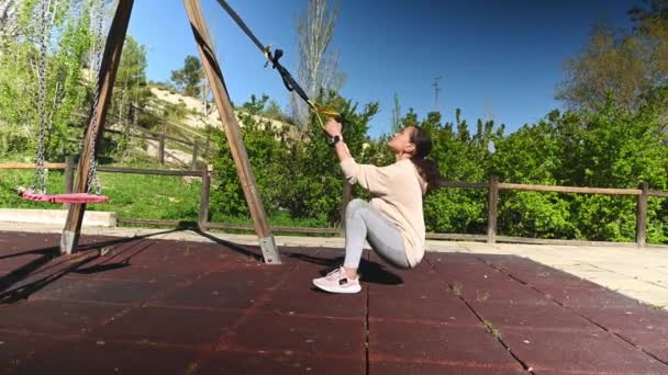 Side view woman squatting, training legs muscles, working out with suspension straps in outdoors sportsground. Determined athlete doing squats, bodyweight training for burning calories and weight loss - Footage, Video