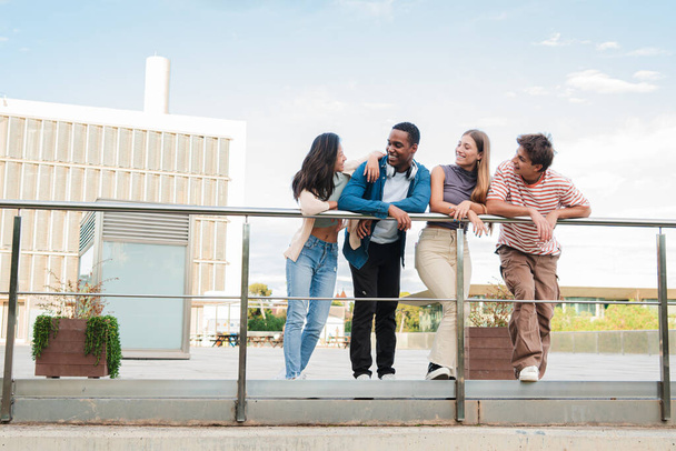 Retrato completo de cuatro estudiantes adolescentes reales sonriendo y hablando juntos en el campus universitario. Grupo de jóvenes amigos conversando sobre un encuentro social. Personas multirraciales charlando - Foto, imagen