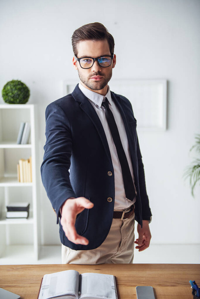 Красивий бізнесмен в окулярах розтягує руку і дивиться на камеру, стоячи на своєму робочому місці в офісі
 - Фото, зображення