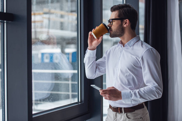 Handsome businessman in eyeglasses is holding a smart phone and drinking coffee while looking out the window in office - Photo, Image