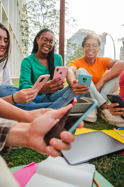 Verticale. Groupe d'étudiants assis sur la pelouse du campus universitaire, s'amusant à utiliser leurs téléphones cellulaires ensemble. Les adolescents partagent des vidéos sur les médias sociaux avec un smartphone. Amis avec téléphone - Photo, image