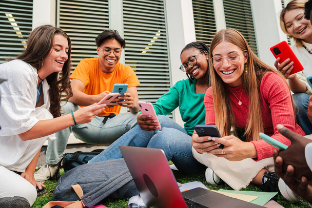 Ομάδα φοιτητών που κάθονται στο γκαζόν της πανεπιστημιούπολης, διασκεδάζοντας χρησιμοποιώντας τα κινητά τους. Έφηβοι άνθρωποι μοιράζονται βίντεο στα μέσα κοινωνικής δικτύωσης με ένα smartphone. Νέοι φίλοι με τηλέφωνο. Υψηλή - Φωτογραφία, εικόνα