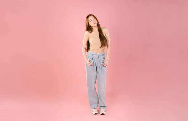 Повне тіло молода усміхнена щаслива азіатська мила жінка 20s в повсякденному джинсі і дивиться на камеру з копіювальним простором ізольовано на рожевому пастельному кольоровому фоні - Фото, зображення