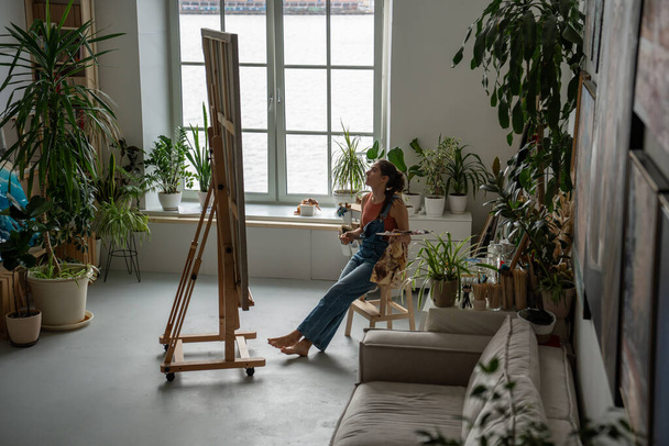Satisfied artistic woman sitting relaxed on chair at easel holding brushes and palette with paints examining huge canvas with drawing. Female painter resting in middle of workflow, taking short break. - Photo, Image