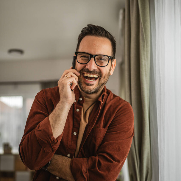 Portret van volwassen man staan terwijl praten op mobiele telefoon en glimlach thuis blij, ontspannen - Foto, afbeelding