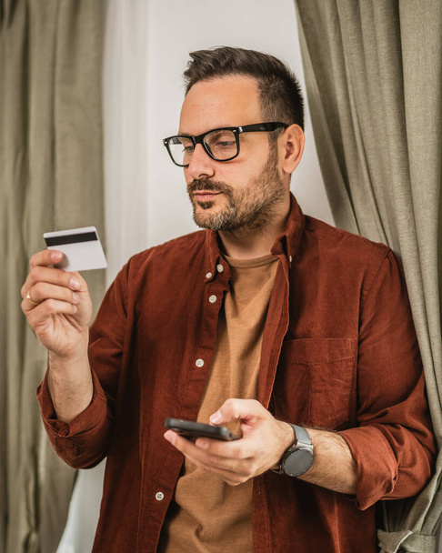 Ritratto di uomo adulto negozio online con cellulare e tenere la carta di credito a casa - Foto, immagini