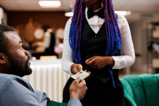 A fiatal afro-amerikai férfi hotel vendége készpénzt ad a pincérnőnek, miközben a hallban pihen, ételt és italt rendel, vendéglátó személyzetnek borravalót ad pihenés közben. Borravalót adó magatartás a vendéglátásban - Fotó, kép