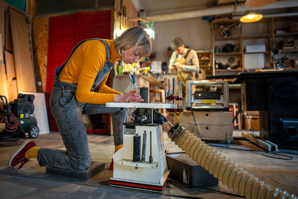 集中した大工の女性は慎重に旋盤に木製品を回します. クラフトスウーマンは注文のための手作りの家具のための特別な機械が付いている木工の仕事項目を磨く家族のジョイナリーの研修会で働きます. - 写真・画像