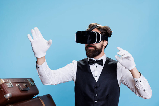 Κομψός γκρουμ που χρησιμοποιεί εικονική πραγματικότητα σε ακουστικά, συμβολίζοντας τη σύγχρονη βιομηχανία φιλοξενίας στο στούντιο. Hotel porter φορώντας λευκά γάντια και vr γυαλιά, παρέχοντας υπηρεσίες πολυτελείας. - Φωτογραφία, εικόνα