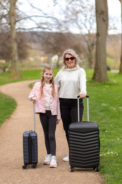 Femme et fille souriantes et posant avec des valises sur fond de parc printanier - Photo, image