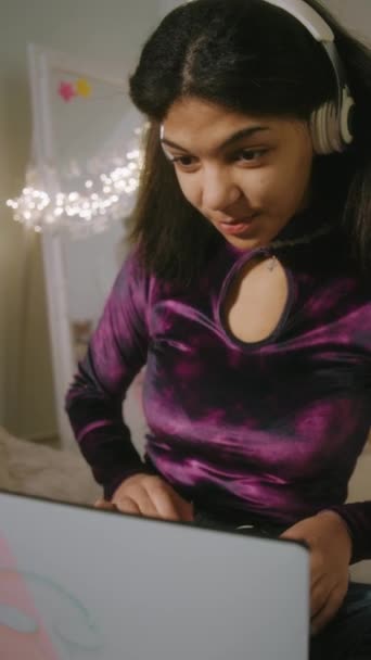 Verticale weergave van tienermeisje in hoofdtelefoon typen op toetsenbord en chatten met een vriend online op laptop zitten in haar gezellige slaapkamer. Afro-Amerikaans meisje besteden vrije tijd en plezier thuis. - Video