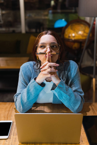 Alto ángulo de la joven mujer de negocios en gafas que sostienen la pluma bajo la nariz y tocar los labios mientras está sentado en la mesa con el ordenador portátil y reflexionar sobre los datos en la noche - Foto, imagen