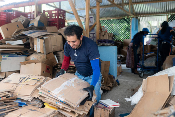 Sonriente joven trabajador hispano que clasifica cartón con compañeros de equipo en un esfuerzo de colaboración en una planta de reciclaje - Foto, imagen