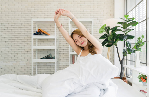 Jonge Aziatische vrouw in wit ondershirt opstaan stretching ontspannen in bed in een lichte, gezellige kamer thuis. Levensstijl concept - Foto, afbeelding