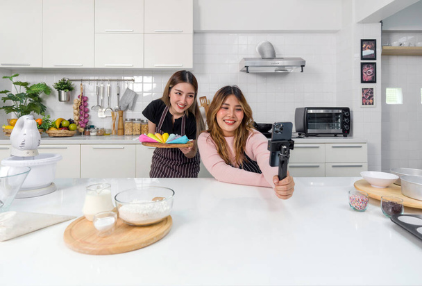 Zwei Frauen vergnügen sich beim Kochen oder Backen in einer modernen Küche. Aufnahme von Videos oder Fotos mit dem Smartphone auf einem Selfie-Stick, während man eine Handvoll bunter Backwerkzeuge oder Dekoration in der Hand hält. - Foto, Bild