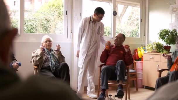 Zpomalený pohyb mladého lékaře mluví s věkem muž cvičit pomocí pedálů na kole a zvedání činky během rehabilitace relace v domově důchodců - Záběry, video