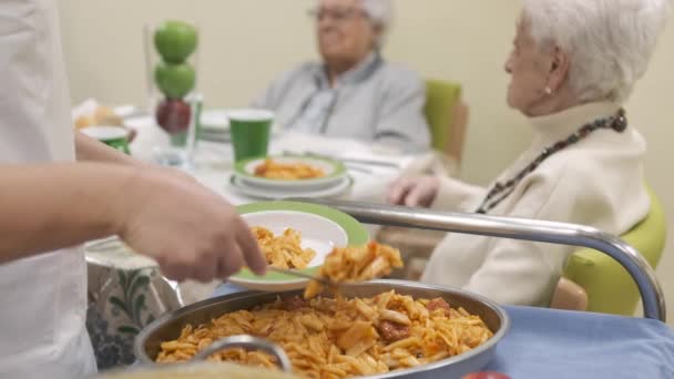 Rack focus slow motion of crop anonymous female worker serving delicious macaroni for aged people gathering around table in canteen of retirement home - Footage, Video