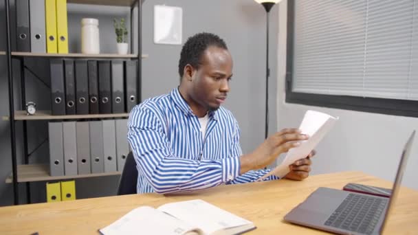 Young focused African employee man with documents, receiving letter, reading legal paper, reviewing project report, sitting at laptop in office - Footage, Video