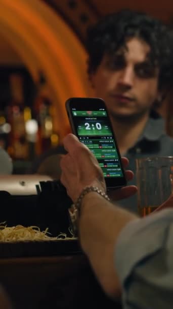 Vertical shot of Mature sports fan bets on football match online using mobile phone, talks with bartender and friend. Man checks game score and bookmaker ratings in app sitting at bar counter in pub - Footage, Video