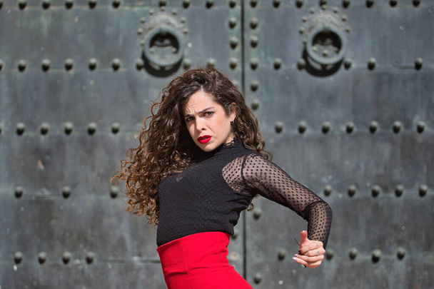 Siyah tişörtlü, kırmızı etekli esmer bir kadının portresi. Arka planda siyah metal kapılı Flamenko dansı yapıyor. Flamenko konsepti, dans, sanat, tipik İspanyolca. - Fotoğraf, Görsel