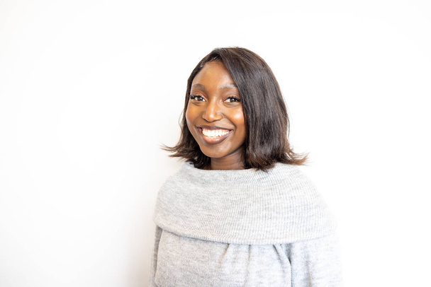 Dit portret vangt een vrolijke jonge zwarte vrouw in een knusse grijze coltrui. Haar stralende glimlach en sprankelende ogen geven een gevoel van geluk en warmte. De schone witte achtergrond - Foto, afbeelding